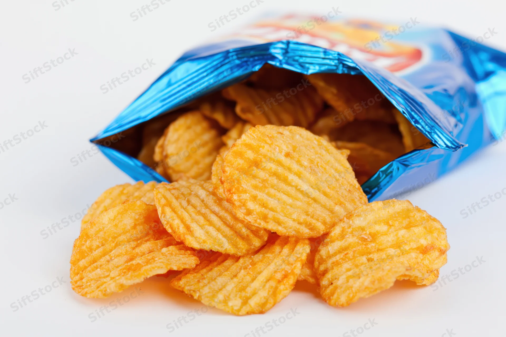 Ridged potato chips spilling from an open blue foil bag on white background