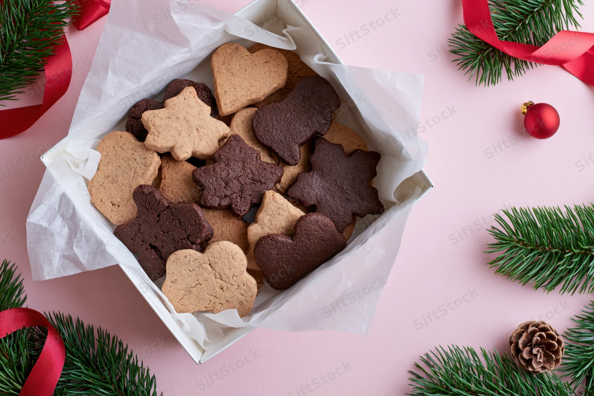 Assorted holiday cookies in a parchment-lined box surrounded by pine branches and red ribbons on a pink surface
