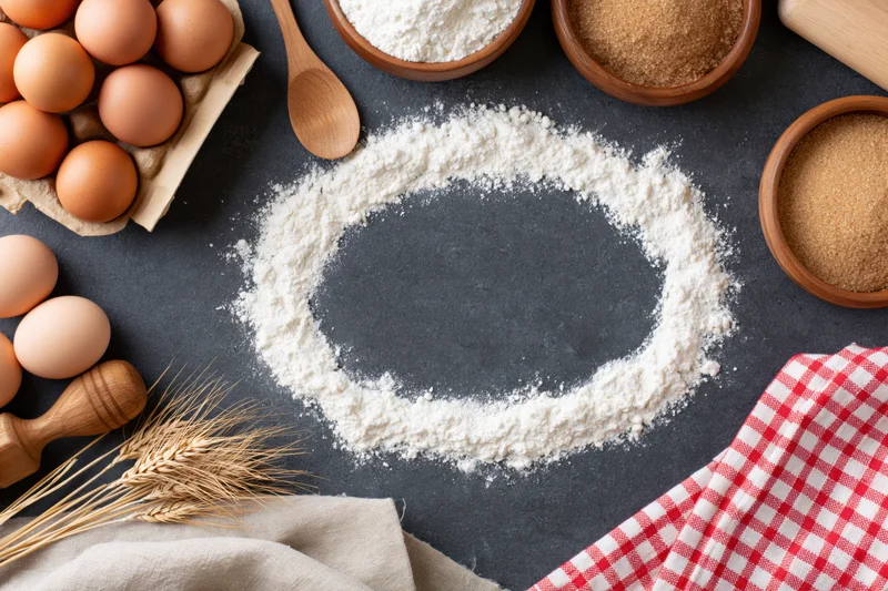 Baking ingredients arranged in a flat lay on a dark stone countertop with flour forming a circle and copyspace in center