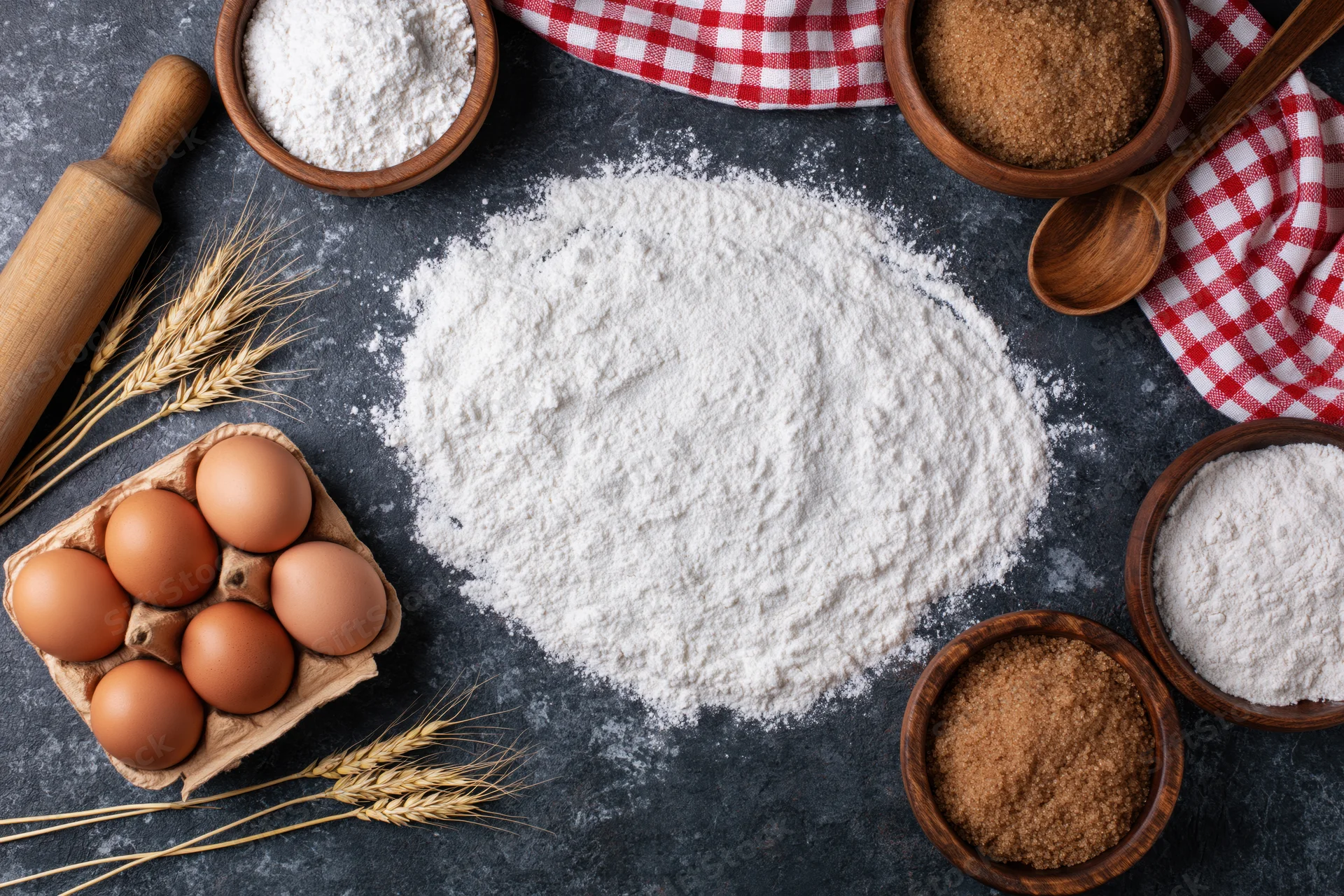 Baking ingredients arranged in a flat lay on a dark stone countertop with flour, eggs, sugar, and wheat stalks