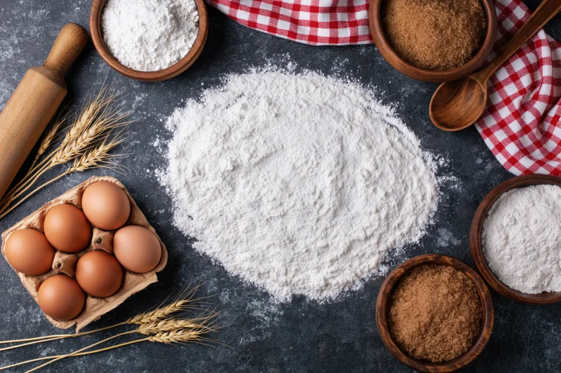 Baking ingredients arranged in a flat lay on a dark stone countertop with flour, eggs, sugar, and wheat stalks