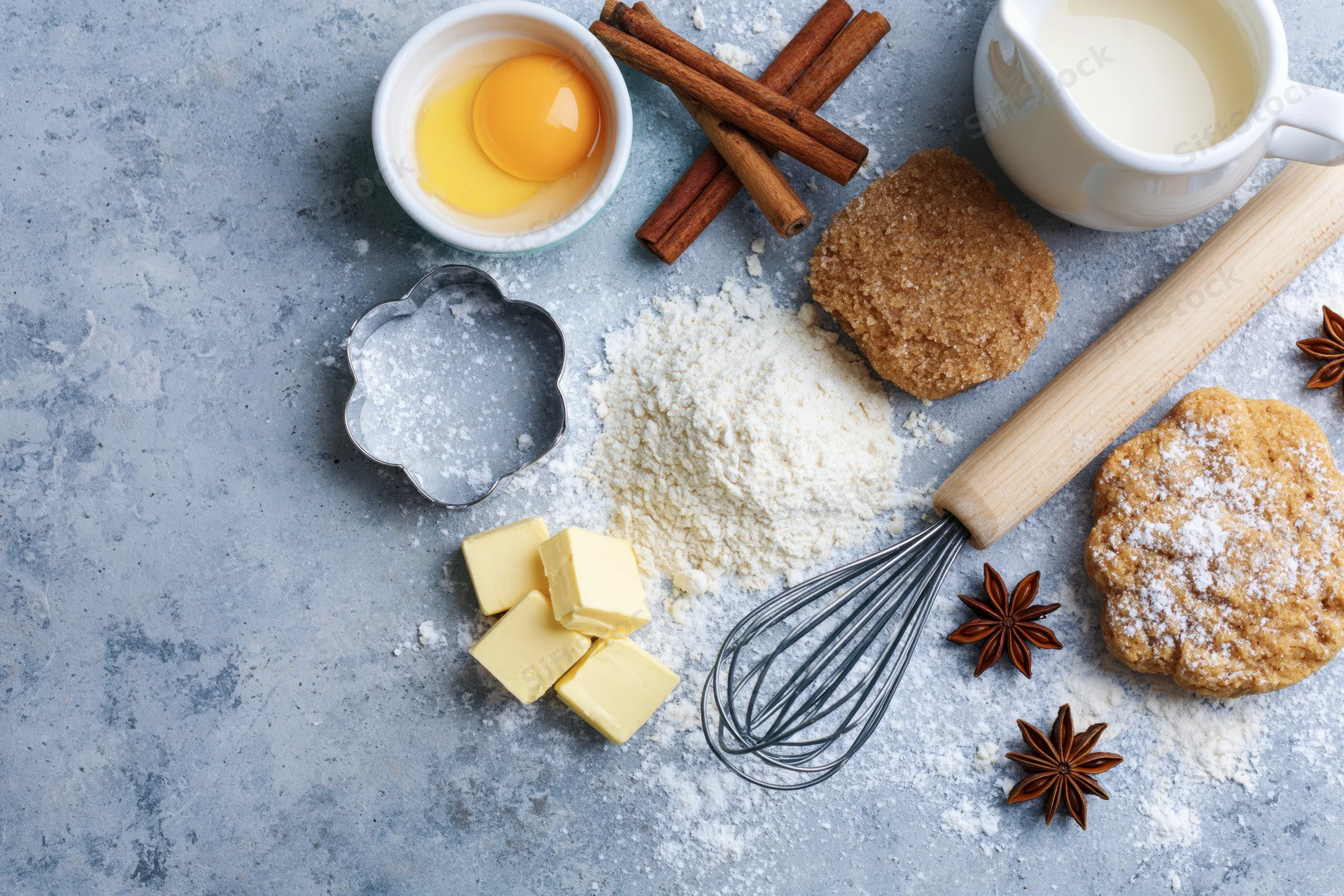 Baking ingredients arranged on a flour-dusted countertop with cookie cutter, whisk and rolling pin