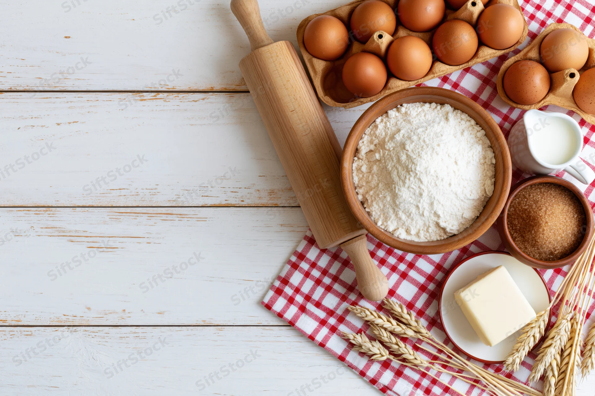 Baking ingredients arranged on a white wooden table with checkered cloth and wheat stalks