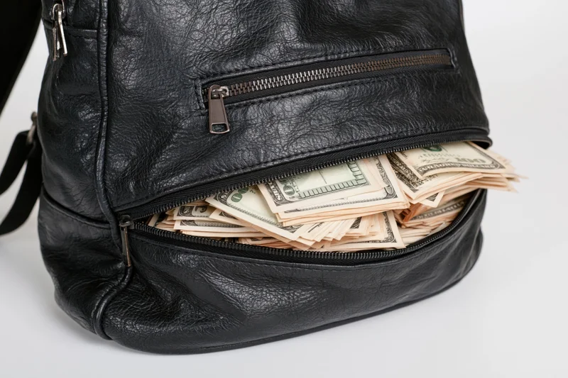 Black leather backpack with open zipper revealing stacks of hundred-dollar bills inside