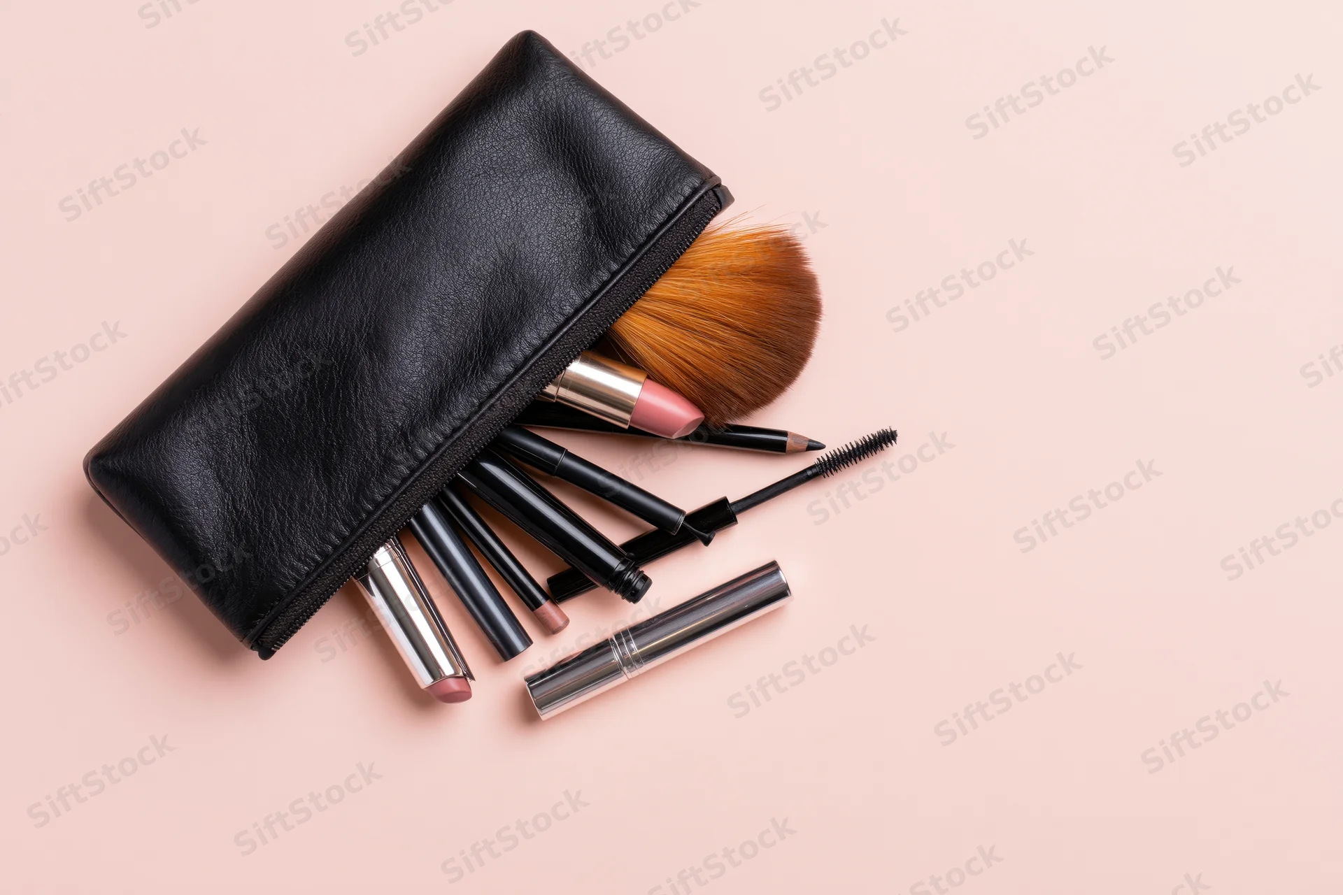 Black leather makeup pouch open with brushes and cosmetics spilling out on pink background