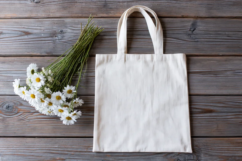 Cream canvas tote bag lying flat next to a loose bunch of daisies on wooden surface