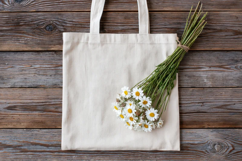 Cream canvas tote bag lying flat on wooden surface with daisies bouquet beside it