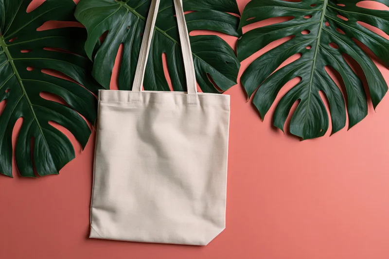 Cream canvas tote bag lying flat on coral surface with large monstera leaves arranged around it