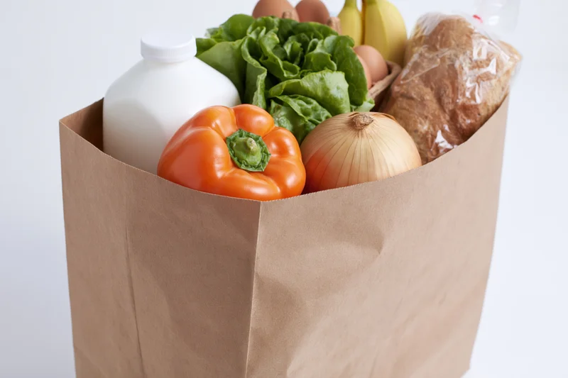 Brown paper grocery bag filled with fresh produce and dairy products on white background