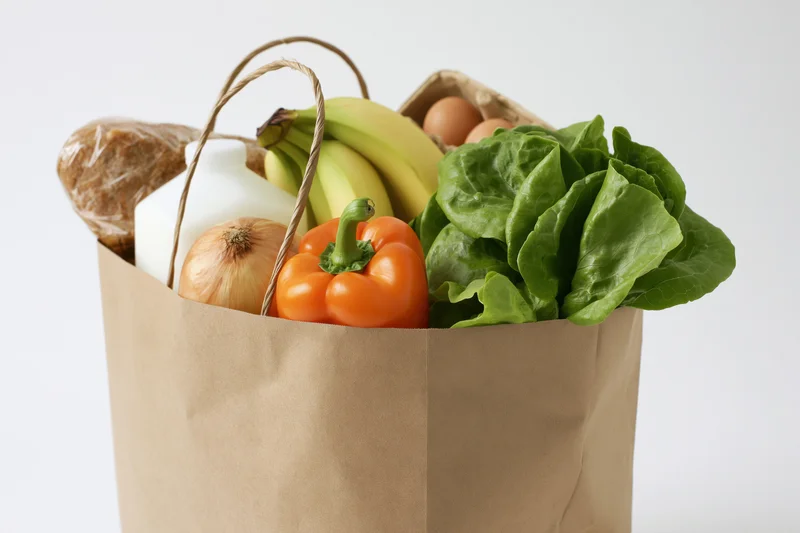 Brown paper bag filled with groceries including vegetables, fruits, milk, bread and eggs against white background