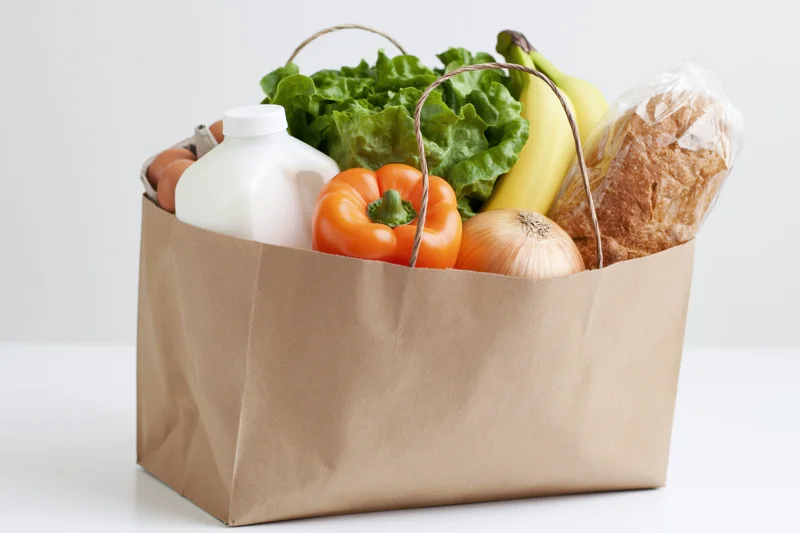 Brown paper grocery bag filled with fresh produce, dairy, bread and eggs on white surface