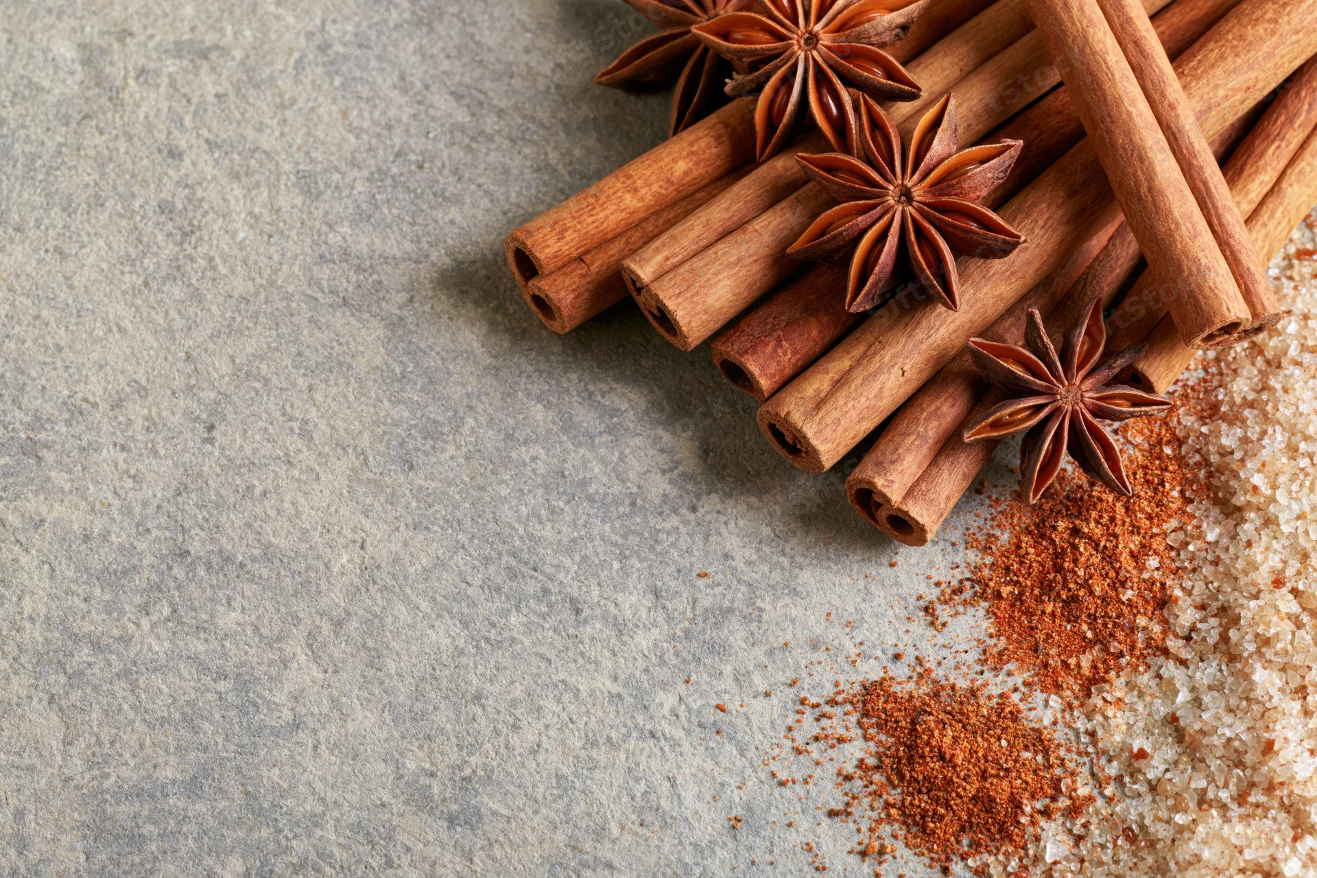 Cinnamon sticks and star anise arranged on a stone surface with ground spice and sugar nearby