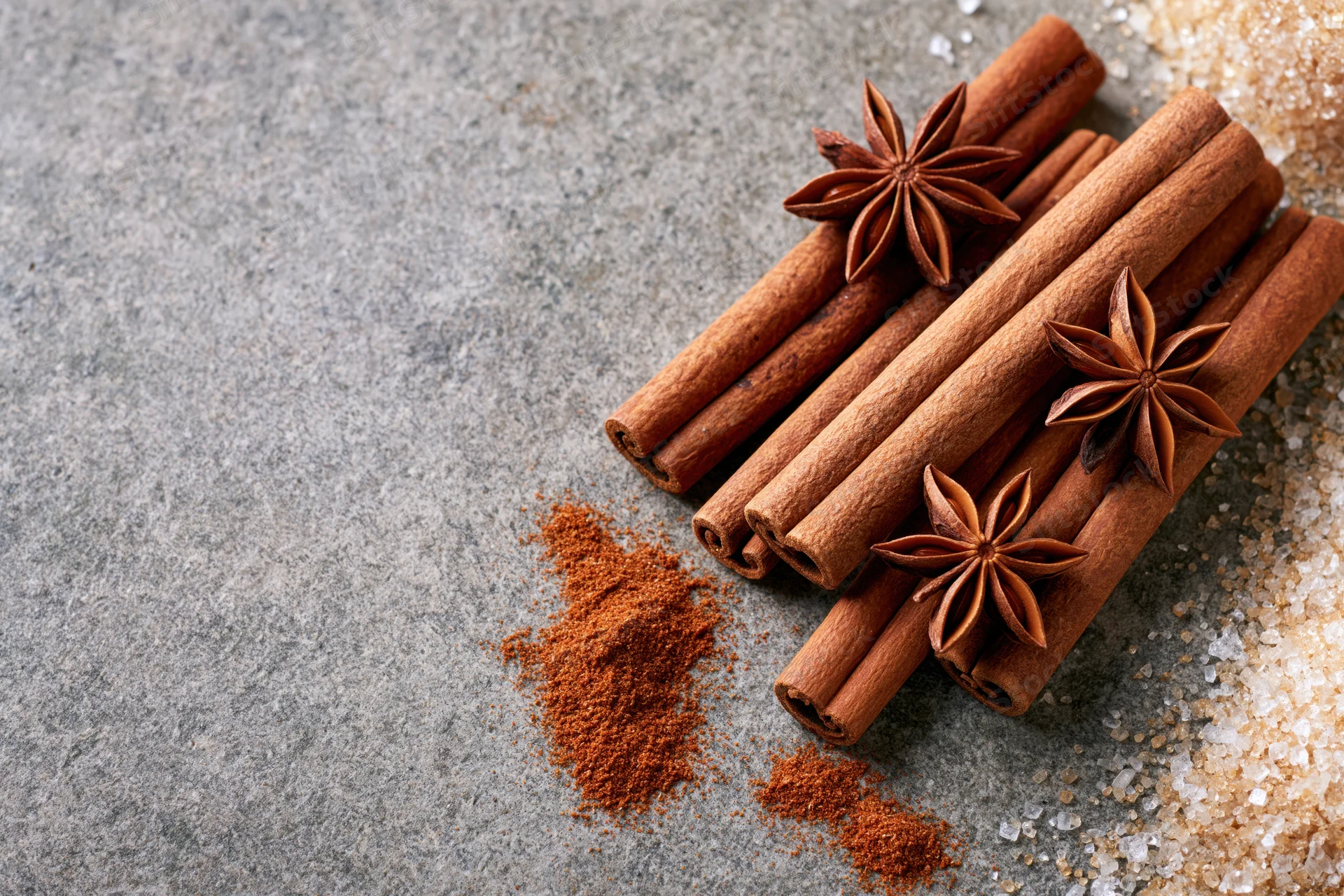 Cinnamon sticks and star anise arranged on a stone surface with ground spice and sugar nearby