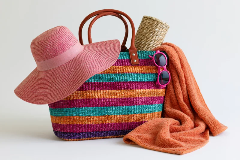 Colorful striped woven straw beach bag with leather handles, pink sunhat, sunglasses and orange towel arranged on white background