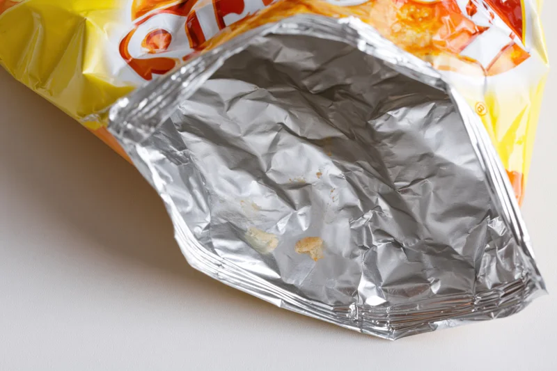 Empty opened chip bag lying on its side showing crumpled aluminum interior with crumbs inside
