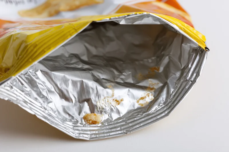 Empty opened chip bag lying on its side showing crumpled aluminum interior with crumbs and residue