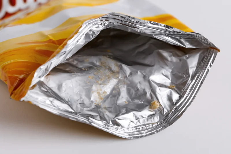 Empty opened chip bag lying on its side showing crumpled aluminum interior with crumbs and residue
