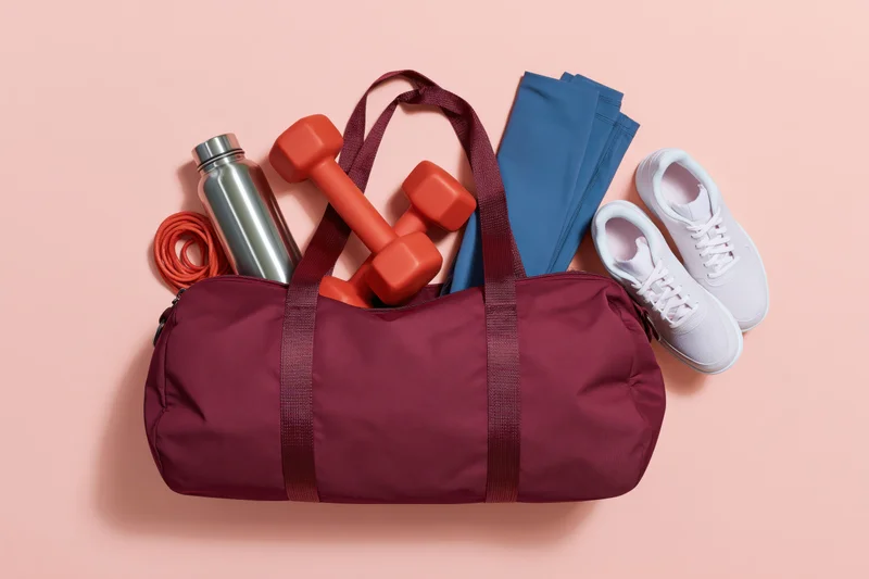 Open burgundy gym duffel bag filled with fitness gear including dumbbells, water bottle, resistance band, leggings and sneakers on a pink background