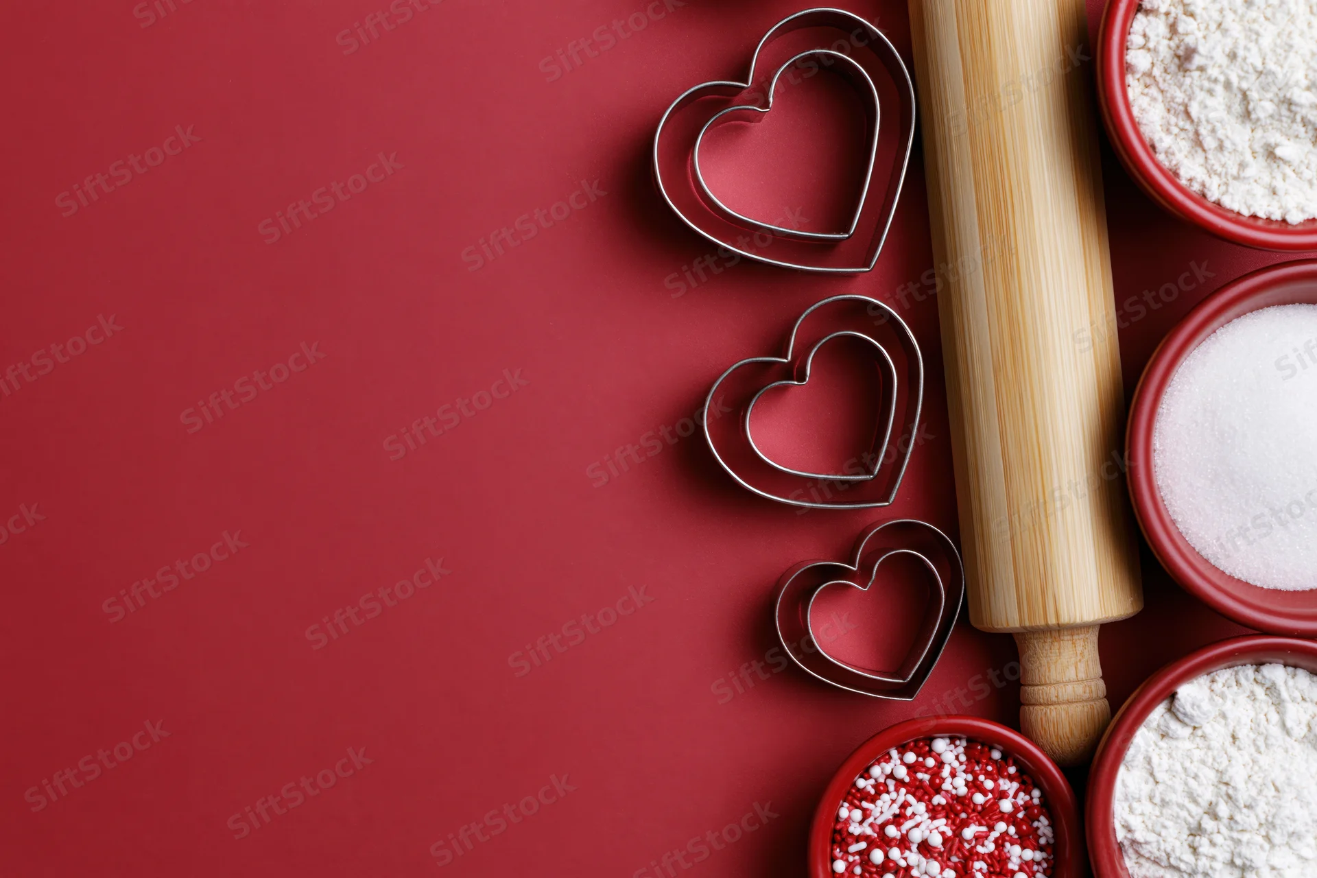 Heart-shaped cookie cutters arranged with baking ingredients on red surface