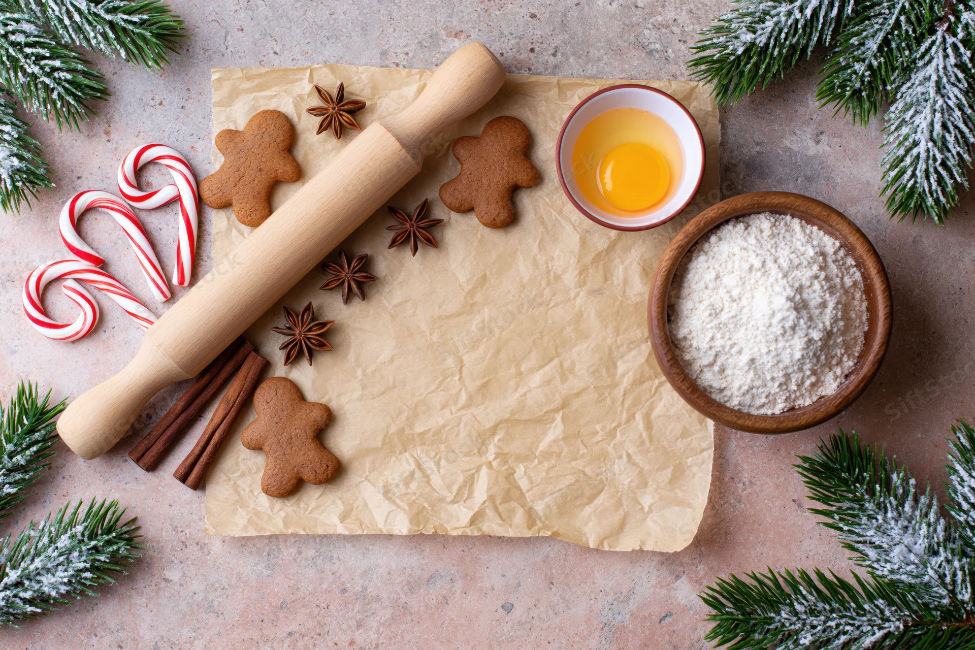 Gingerbread cookies and baking ingredients arranged on parchment with evergreen branches and candy canes for christmas baking