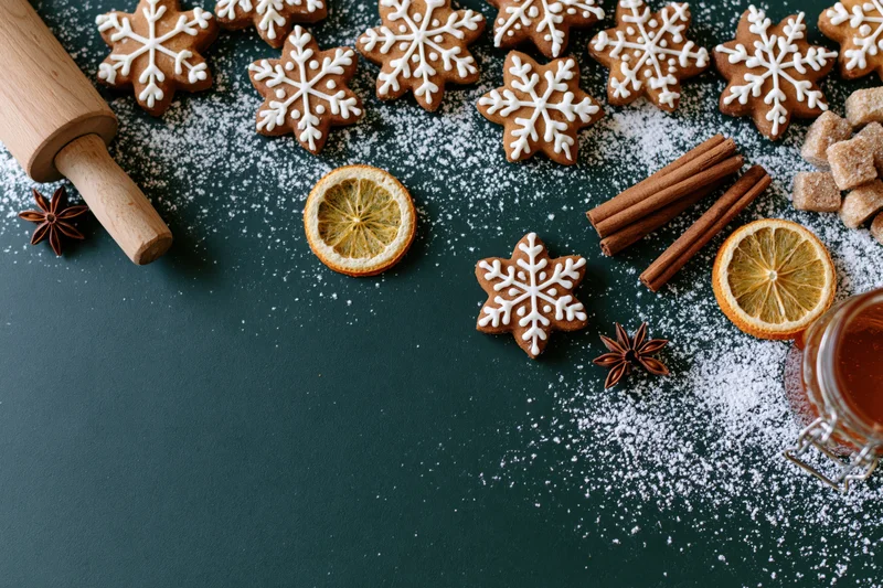 Snowflake-shaped gingerbread cookies with cinnamon sticks, dried orange slices, star anise, powdered sugar, and a rolling pin arranged on a dark green surface in a flat lay composition