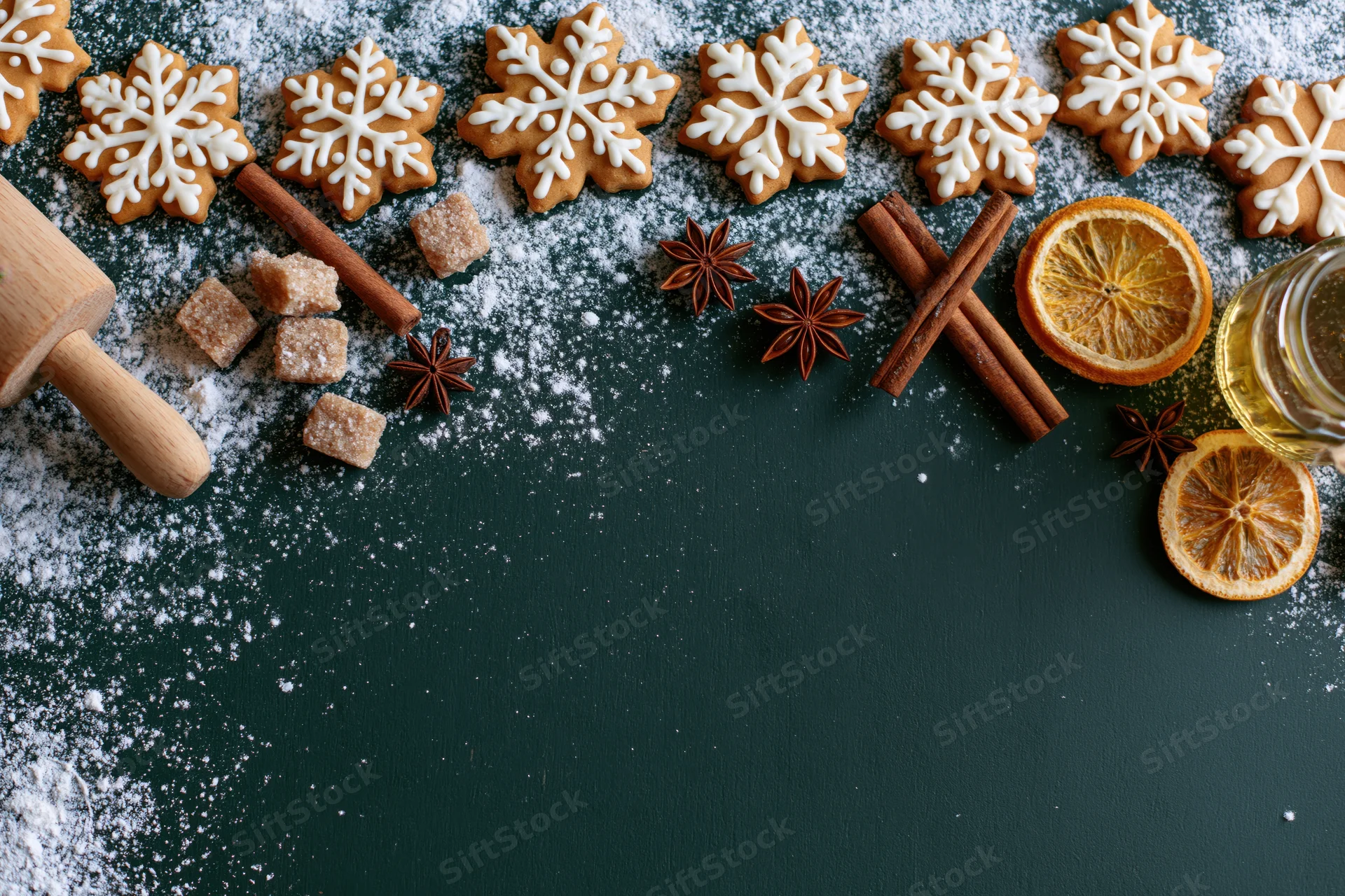 Snowflake-shaped gingerbread cookies with spices and dried orange on dark green surface, flat lay arrangement with copyspace