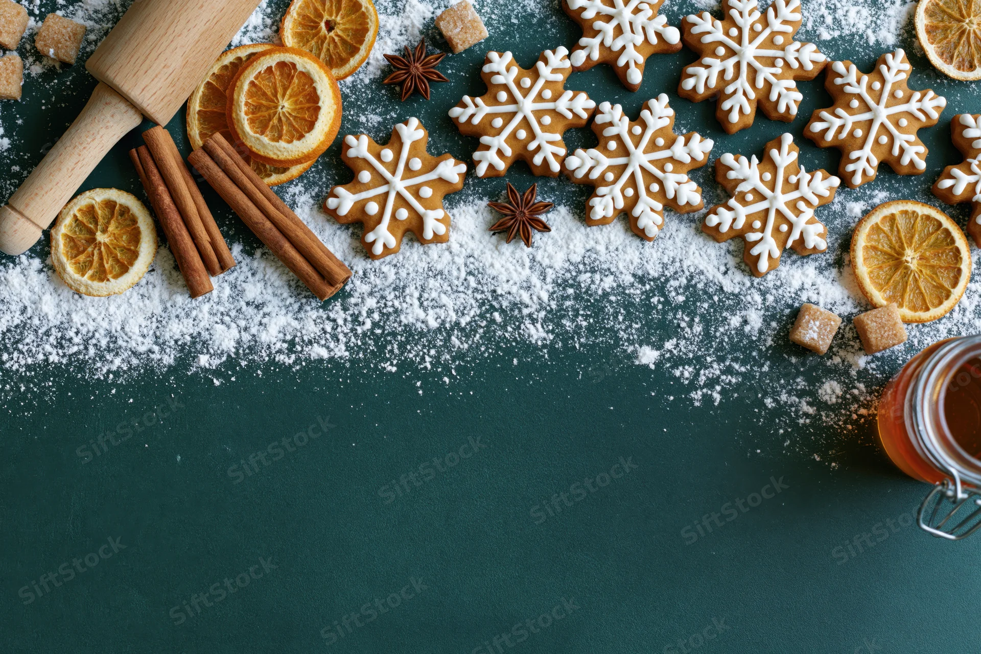 Snowflake-shaped gingerbread cookies with cinnamon sticks, dried oranges, and powdered sugar on a dark green surface, arranged in a flat lay with copyspace below