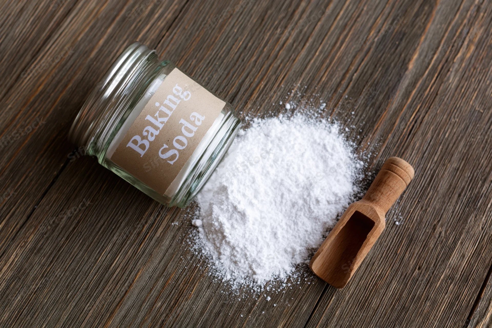 Glass jar labeled baking soda tipped over with white powder spilled onto wooden surface beside wooden scoop