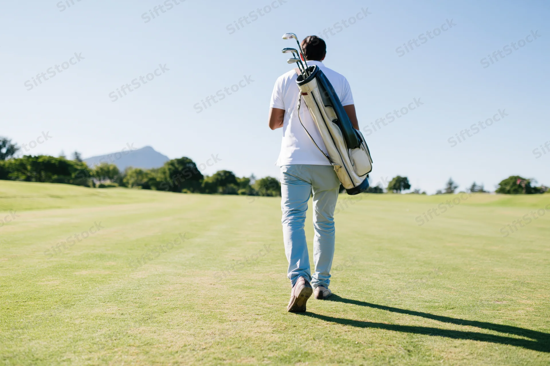 Man walking across a golf course fairway carrying a golf bag with clubs, back view in sunlight