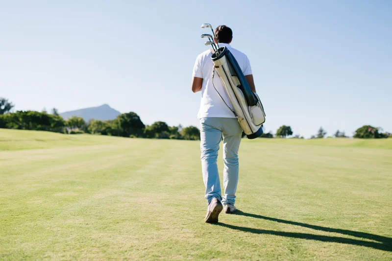 Man walking across a golf course fairway carrying a golf bag with clubs, back view in sunlight