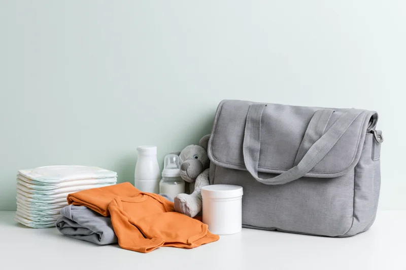Grey diaper bag with baby essentials arranged on a white surface against a mint wall