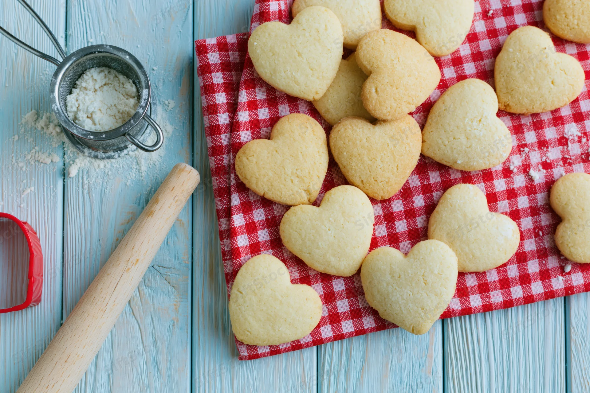 Heart-shaped shortbread cookies on a red gingham cloth beside baking tools on a blue wooden table