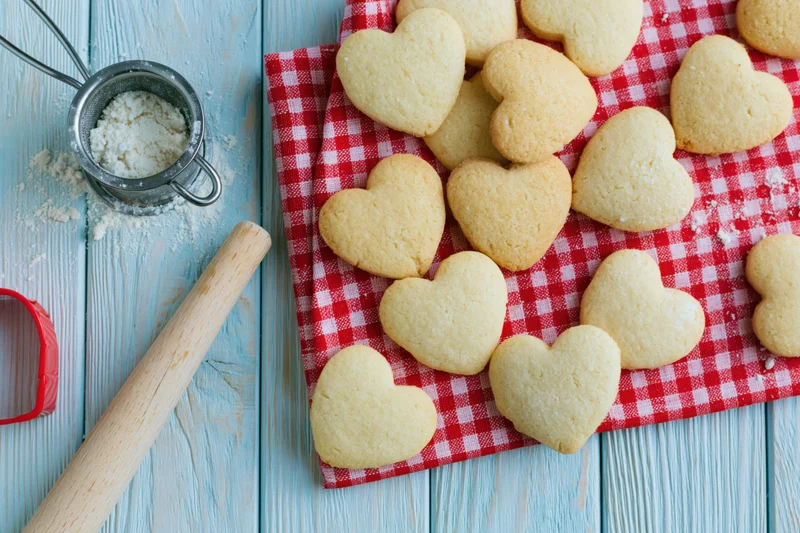 Heart-shaped shortbread cookies on a red gingham cloth beside baking tools on a blue wooden table
