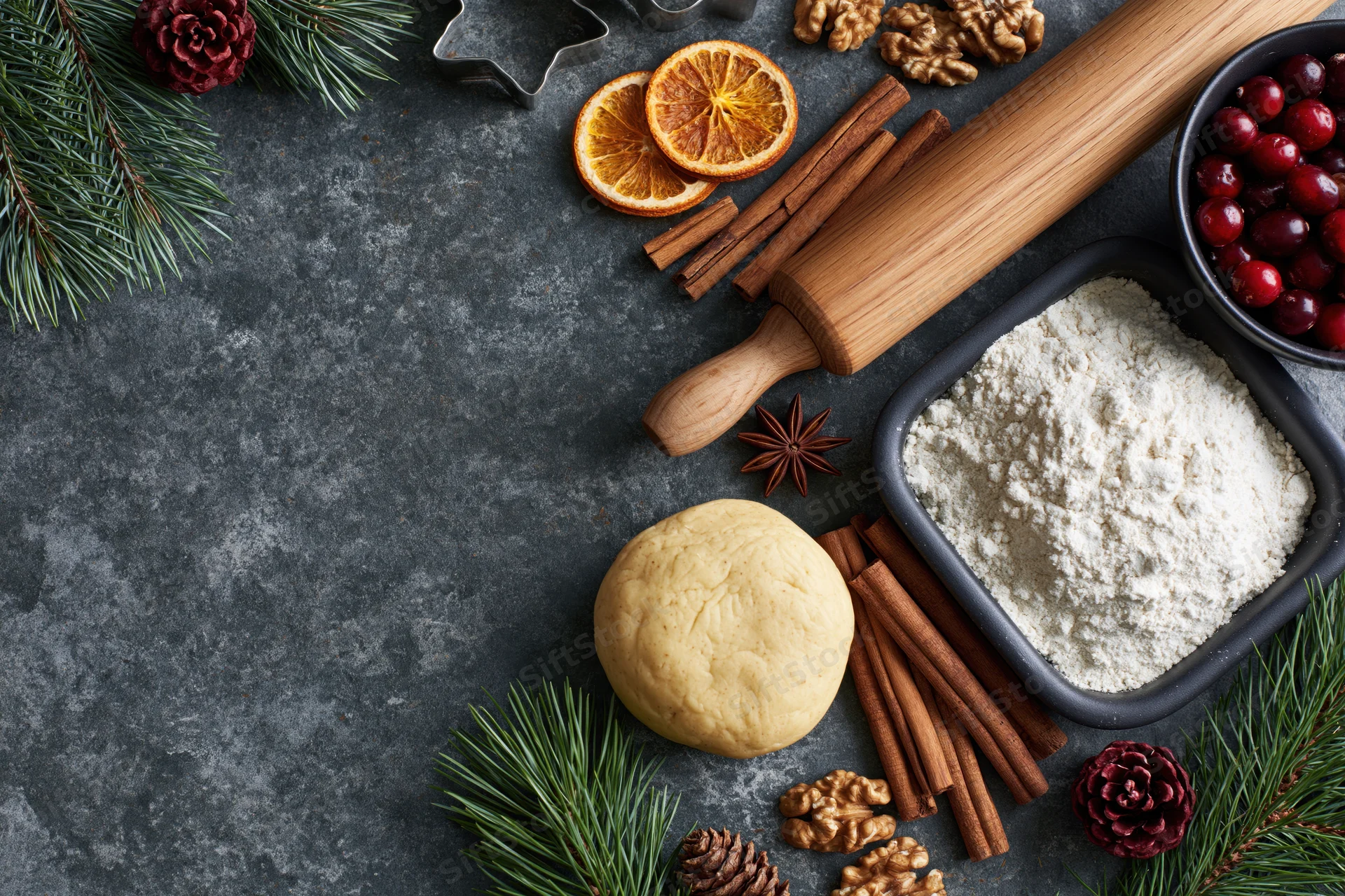 Holiday baking ingredients arranged on dark slate surface with pine and cranberries