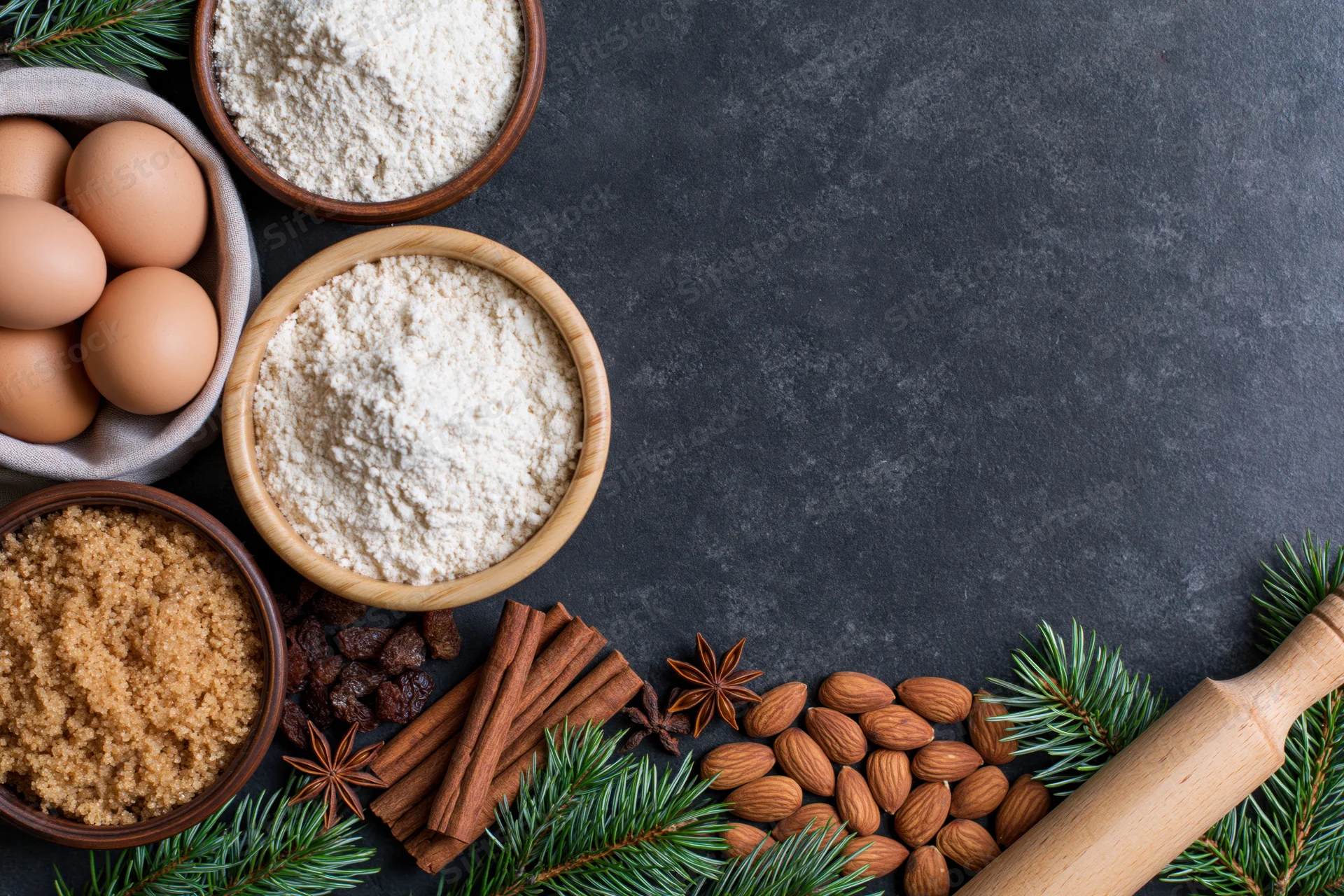 Holiday baking ingredients arranged along the bottom edge of a dark countertop with pine branches and a rolling pin