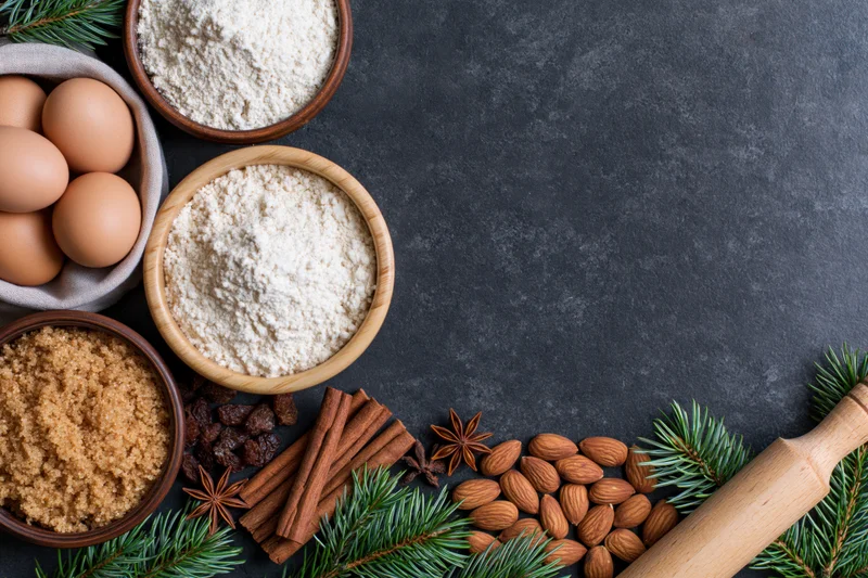 Holiday baking ingredients arranged along the bottom edge of a dark countertop with pine branches and a rolling pin