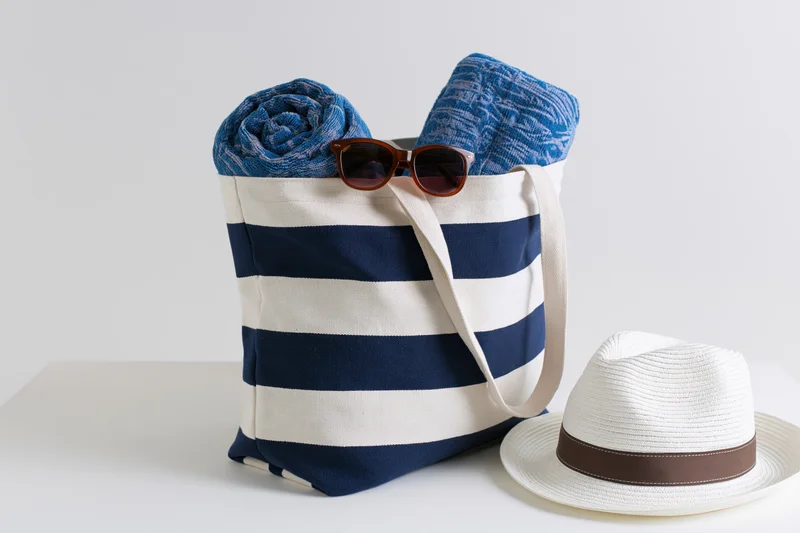 Navy and white striped canvas tote bag filled with rolled towels and sunglasses, beside a straw hat on a white surface