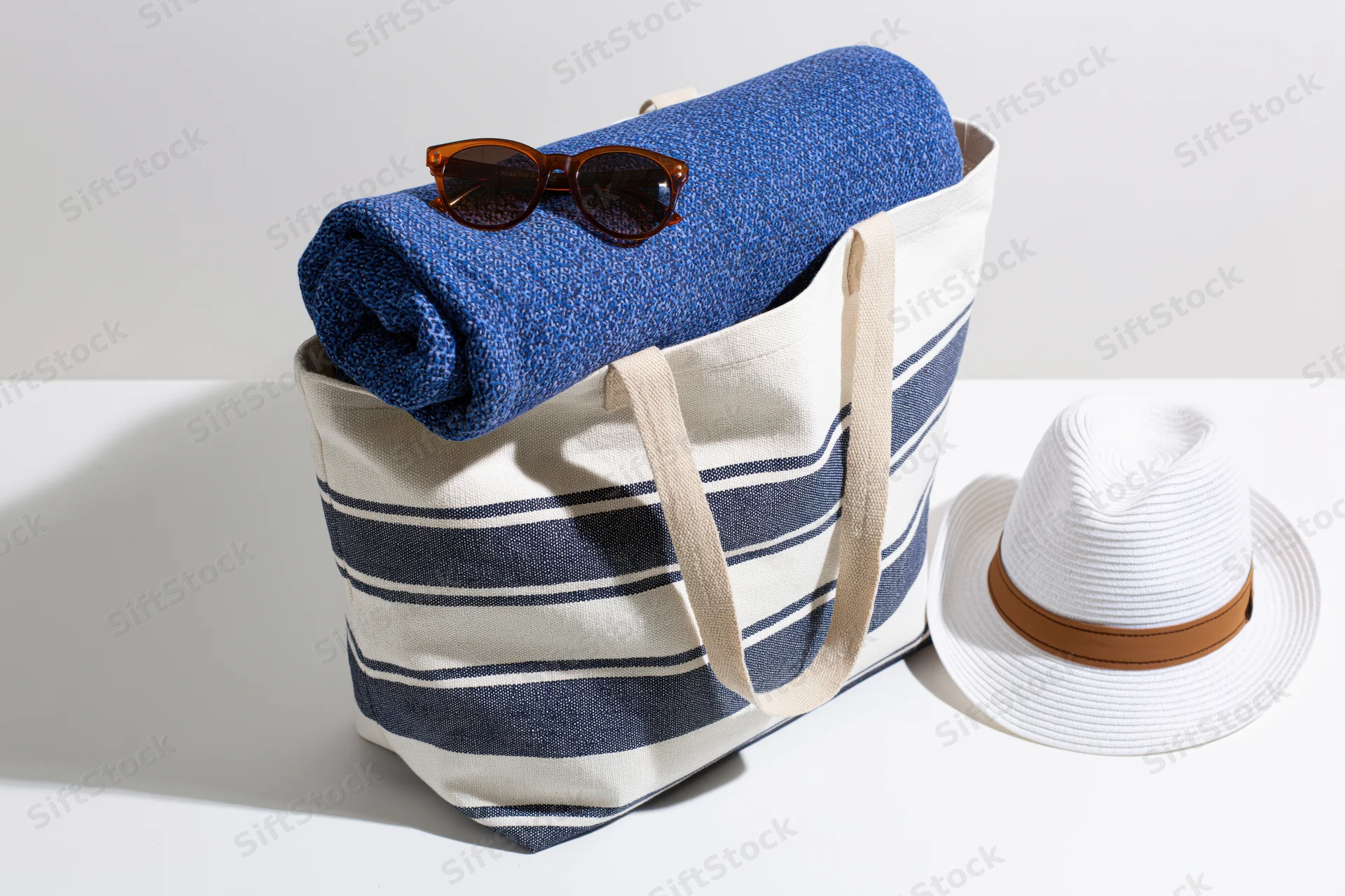 Navy and white striped canvas tote bag holding a rolled blue towel and sunglasses with a white straw hat beside it on a white surface