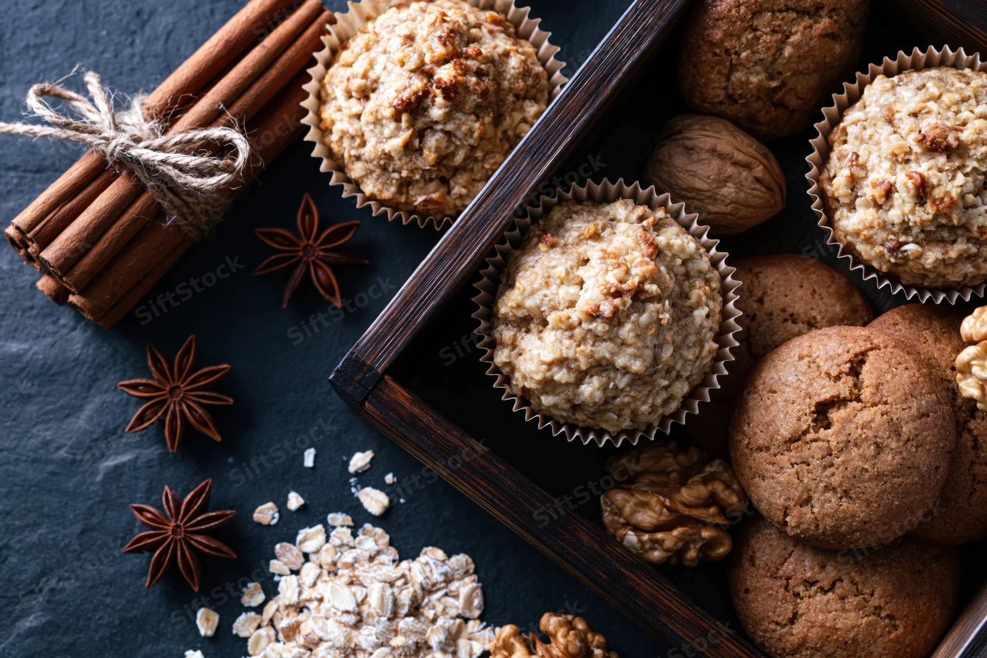 Oat muffins and cookies in paper liners with cinnamon, star anise, walnuts and oats on dark slate surface