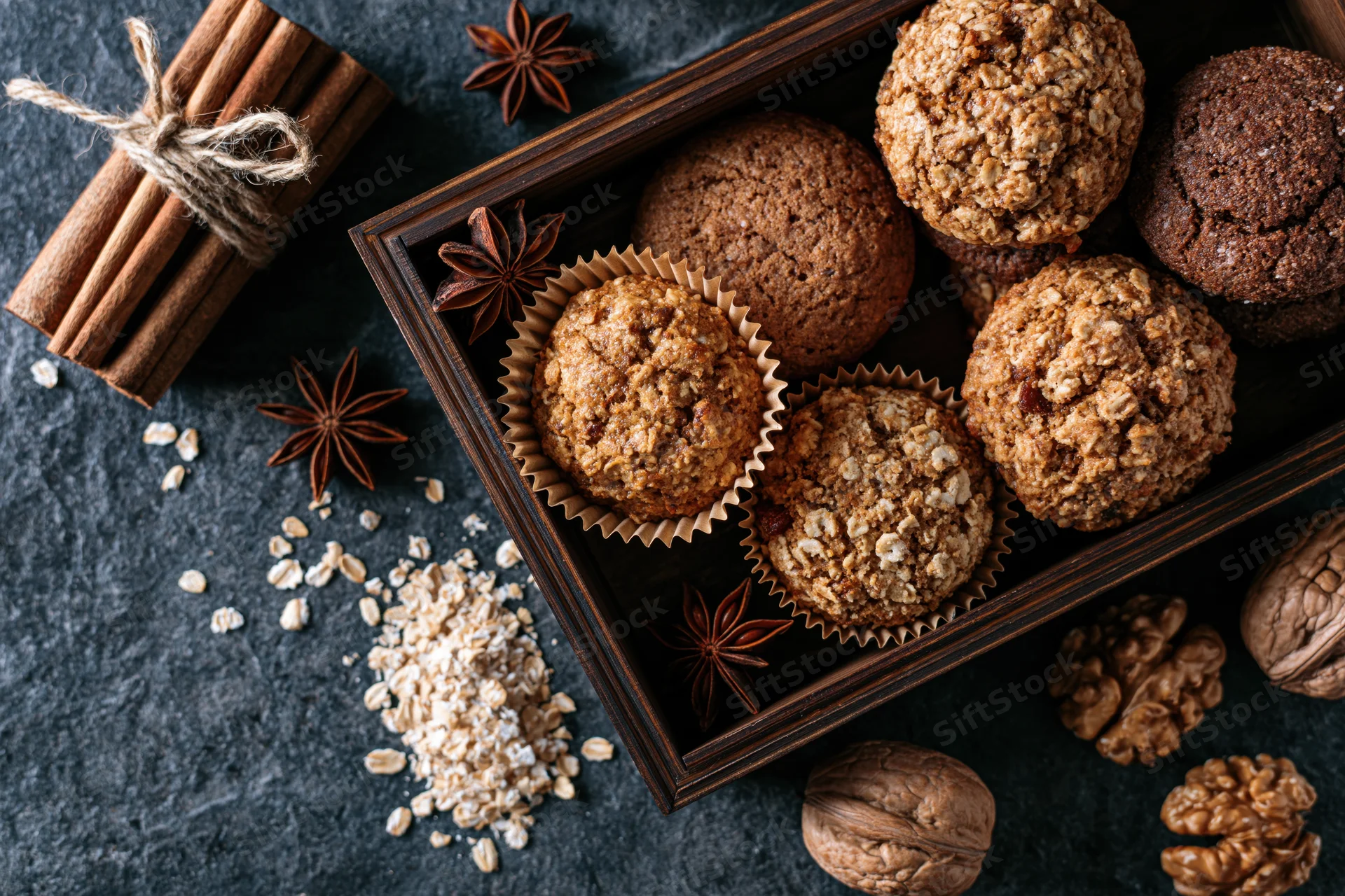 Oat muffins and cookies in paper liners arranged in a dark wooden tray with cinnamon, star anise, oats, and walnuts around