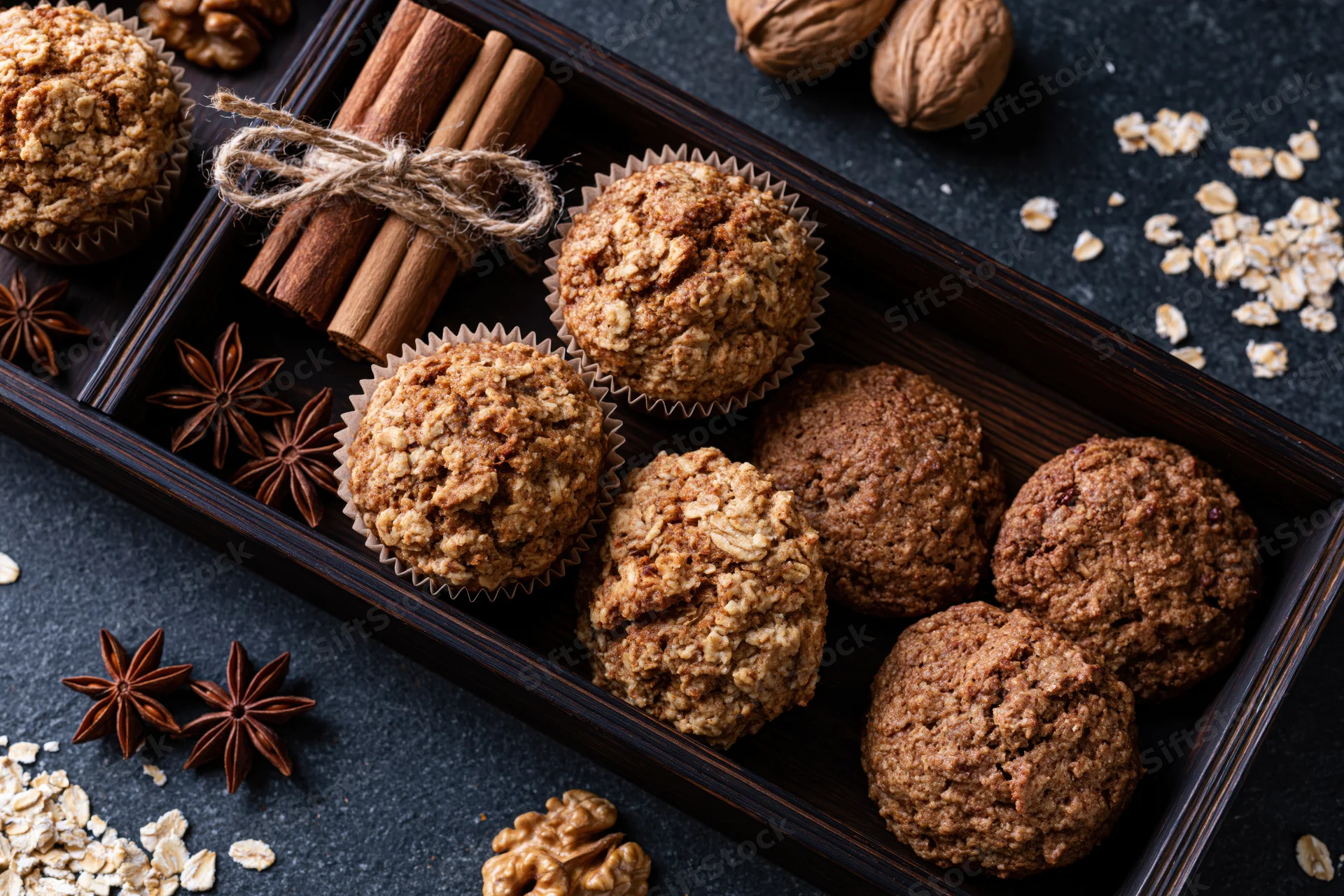 Oat muffins and cookies in paper liners arranged on a dark wooden tray with cinnamon sticks, star anise, and scattered oats