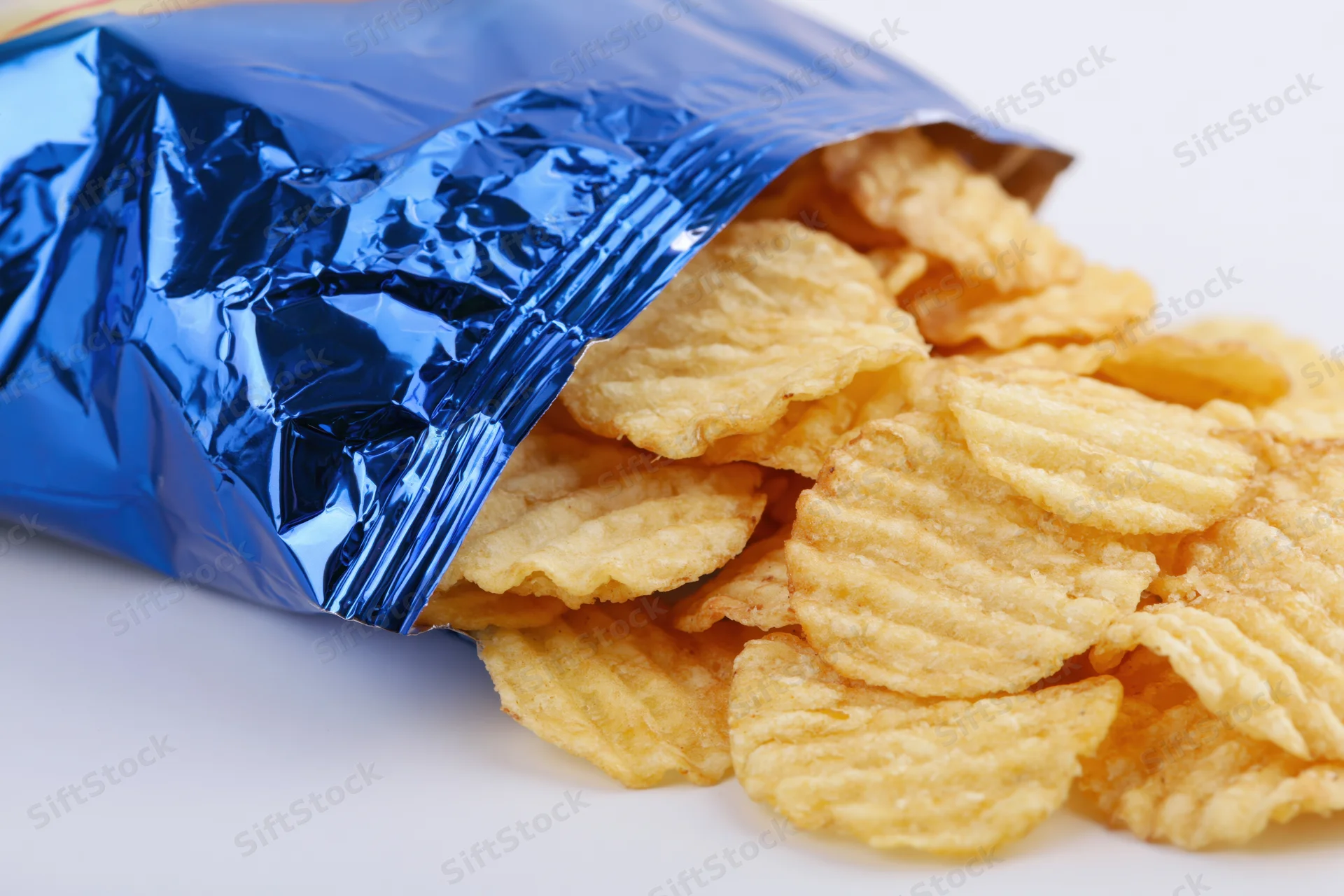 Rippled potato chips spilling from an open blue metallic snack bag on white surface