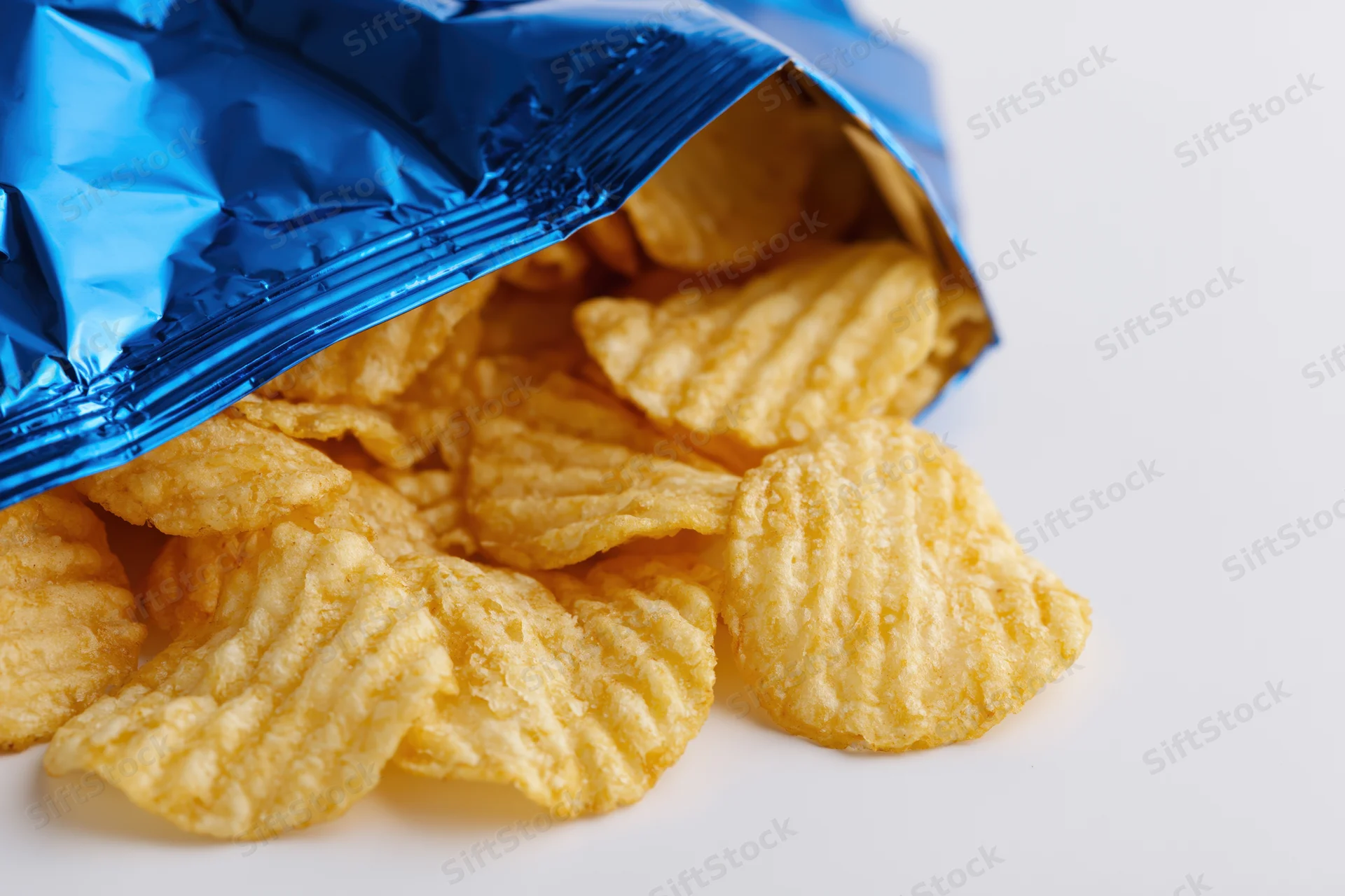 Rippled potato chips spilling from an open blue metallic snack bag on white surface