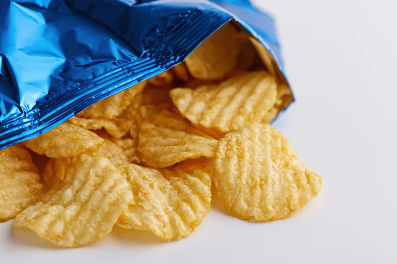 Rippled potato chips spilling from an open blue metallic snack bag on white surface