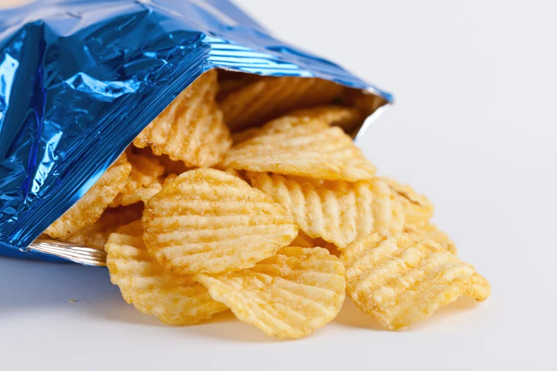 Open blue metallic snack bag tipped on its side with rippled potato chips spilling out onto white surface