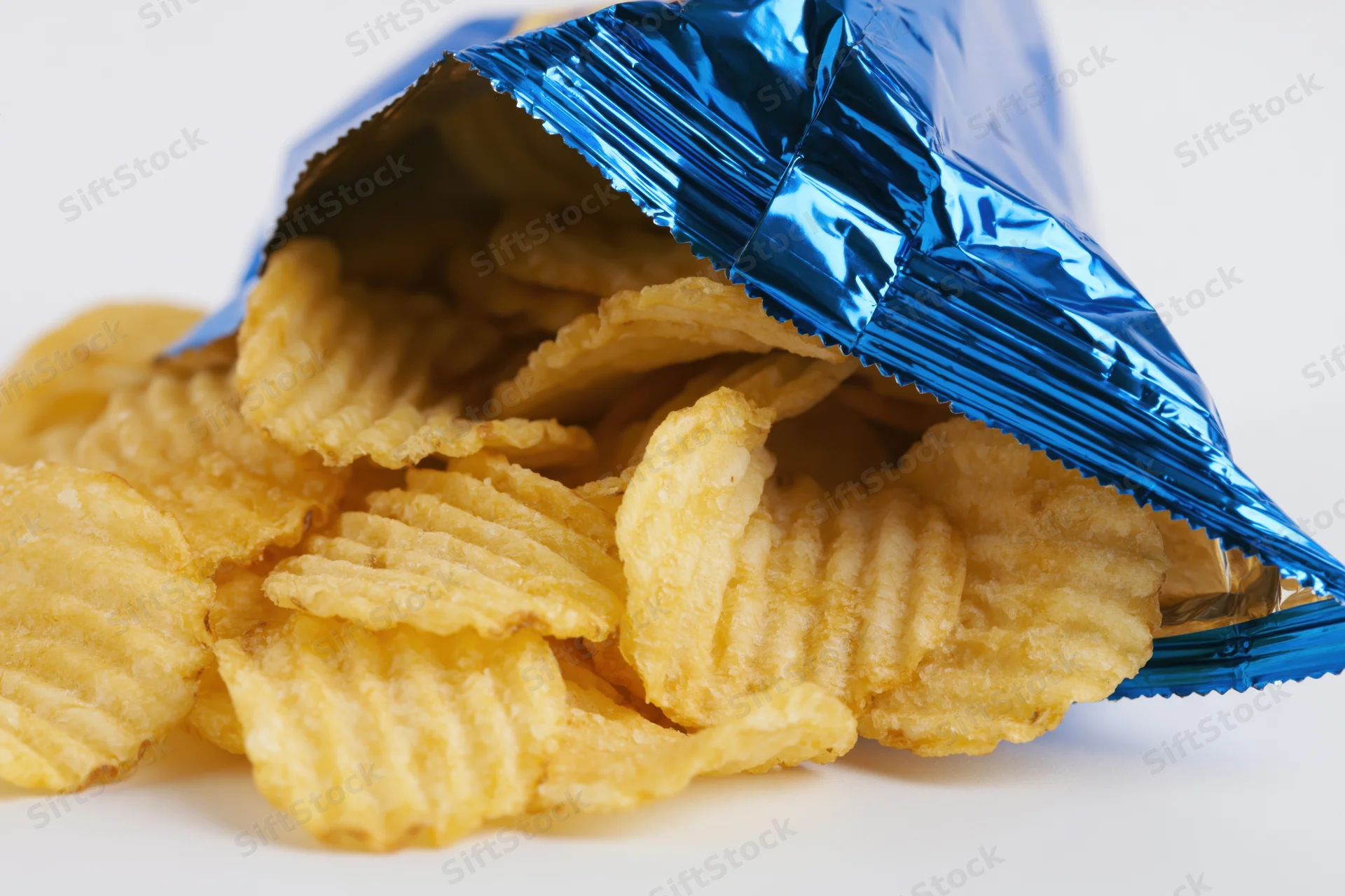 Open blue metallic snack bag tipped on its side with rippled potato chips spilled out onto white surface