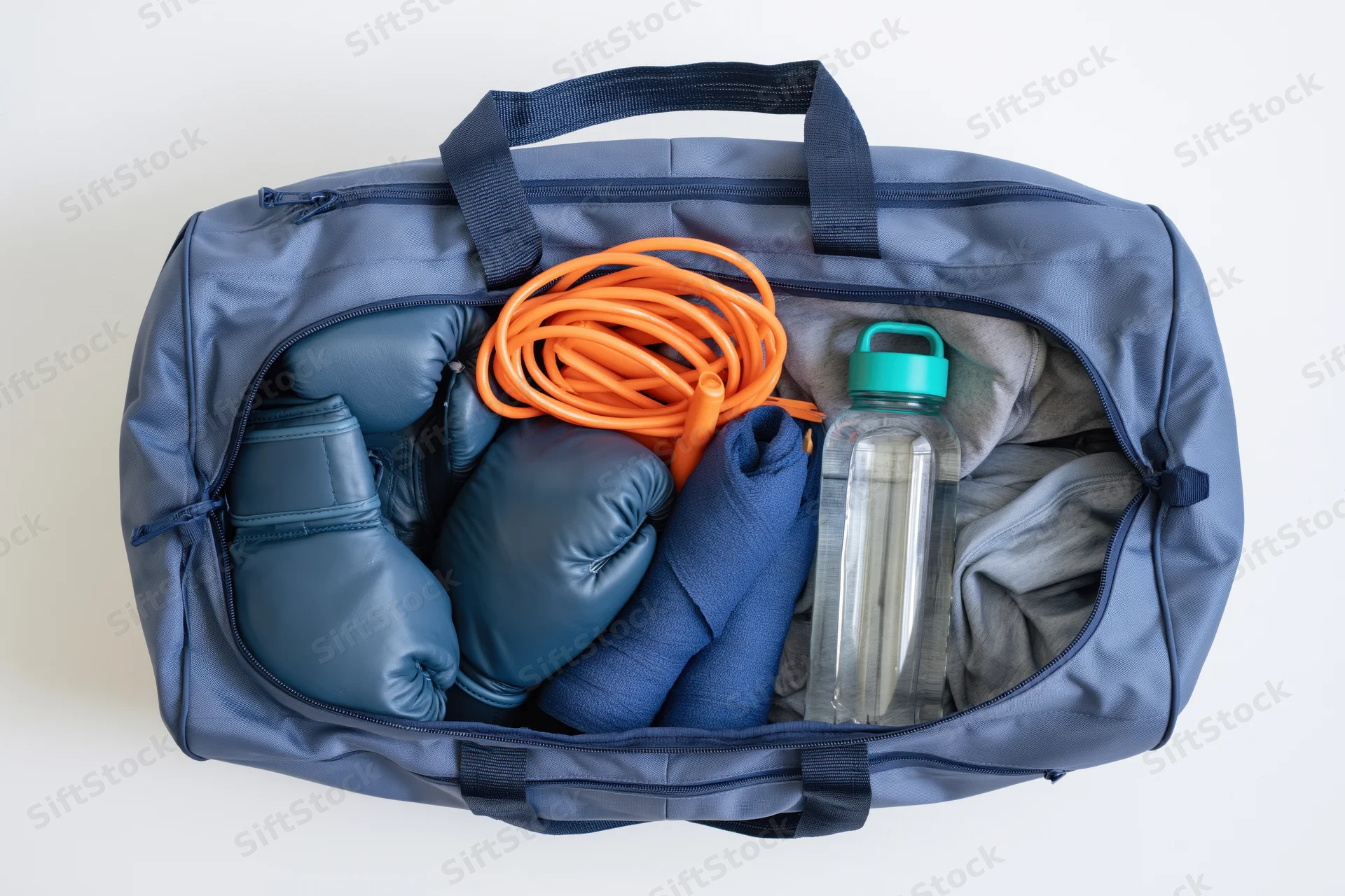 Open blue gym bag filled with boxing gloves, jump rope, water bottle, towel and clothes on white background