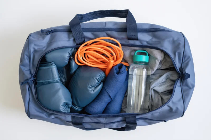 Open blue gym bag filled with boxing gloves, jump rope, water bottle, towel and clothes on white background