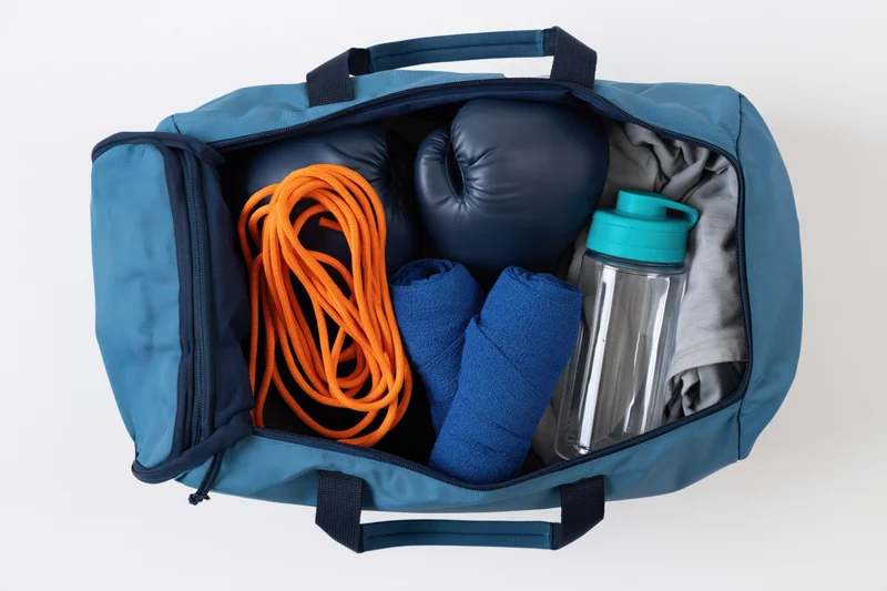 Open blue gym bag filled with boxing gloves, jump rope, water bottle, towels and workout clothes on white background