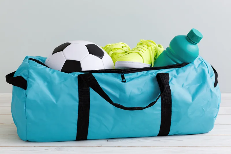 Cyan sports duffel bag filled with soccer ball, lime sneakers and water bottle on white wooden surface